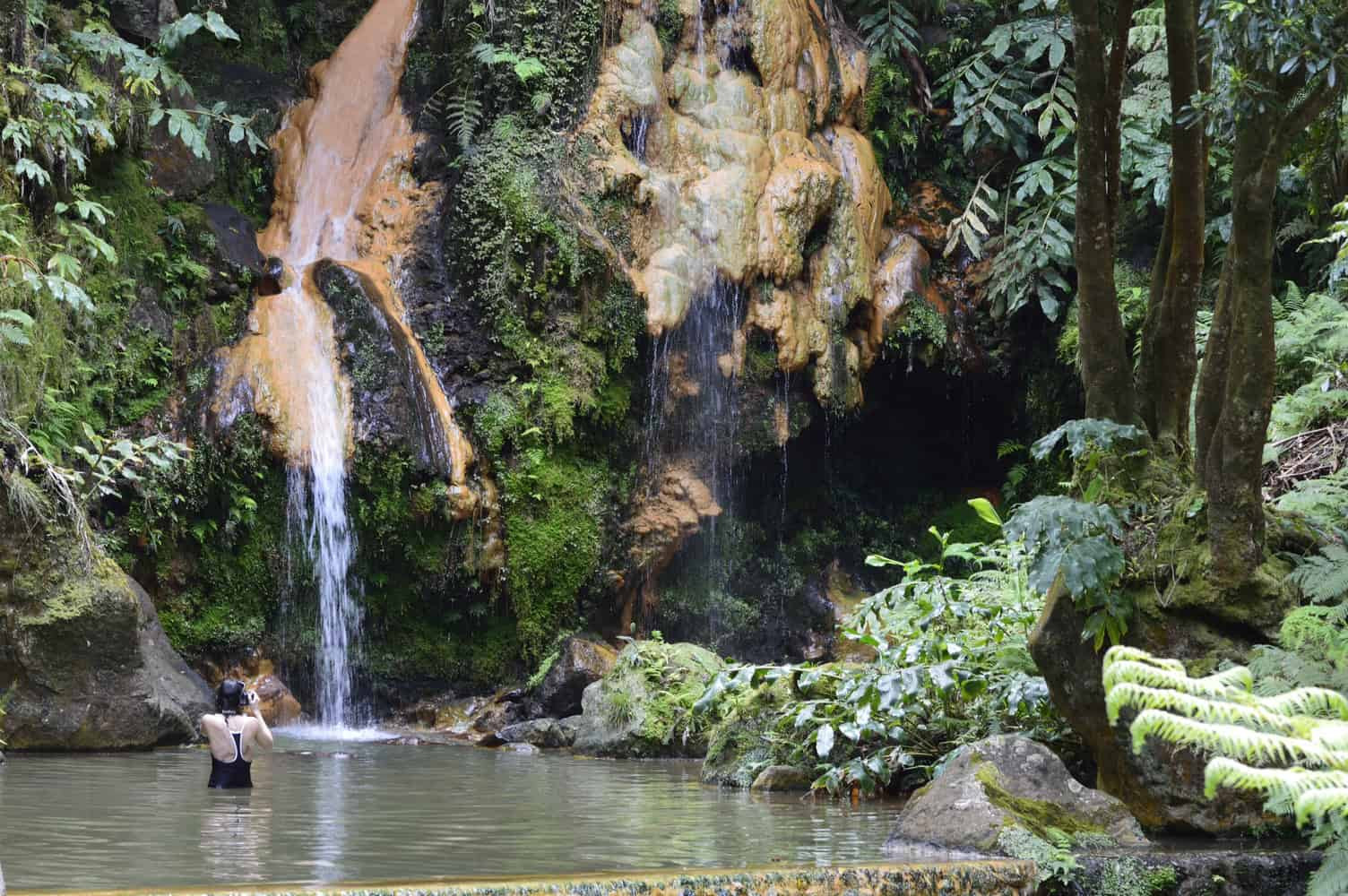 Person bathing in natural hot spring under a waterfall at Caldeira Velha, surrounded by lush green forest on São Miguel Island, Azores